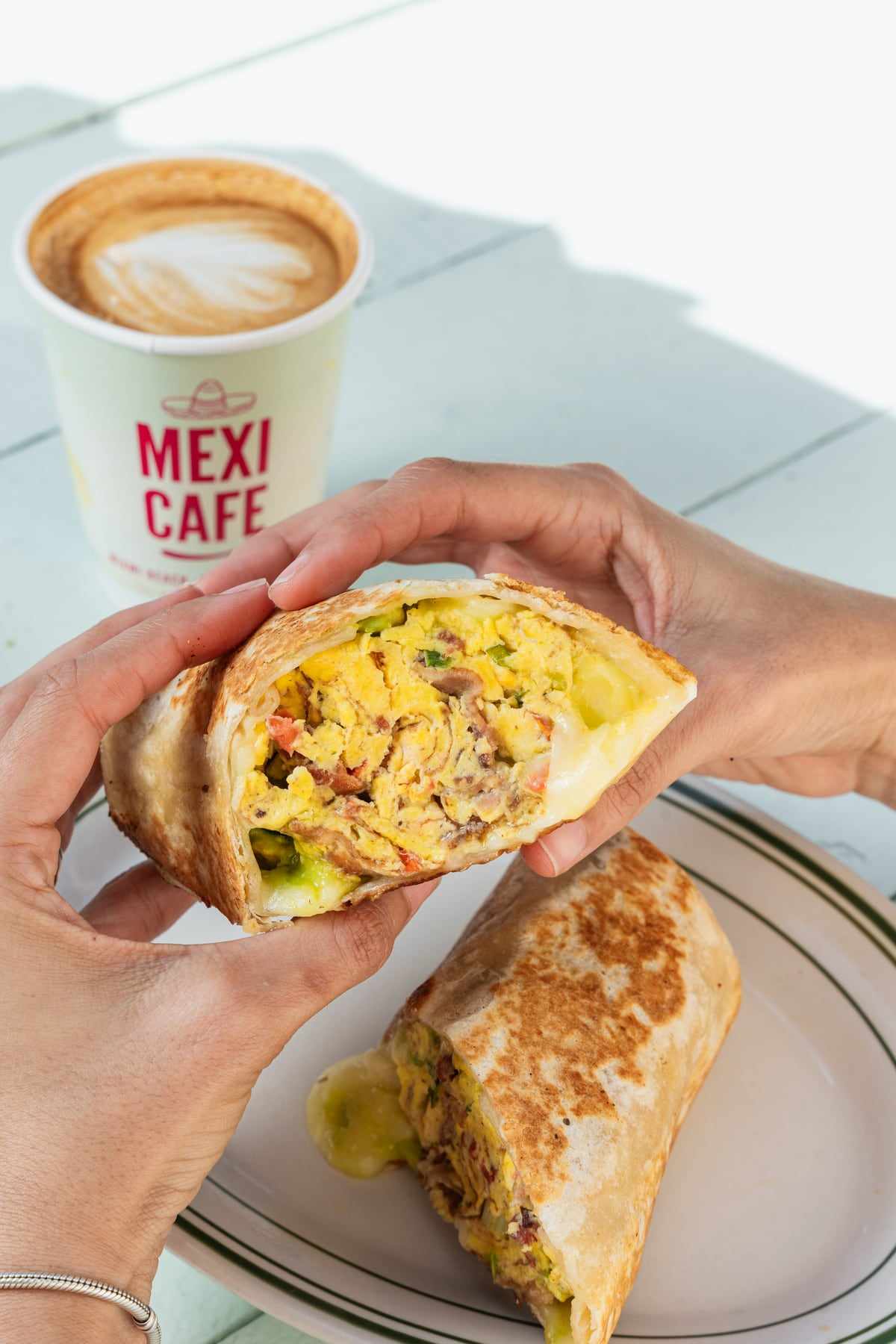 A breakfast burrito held in two hands next to a Mexi Cafe coffee cup