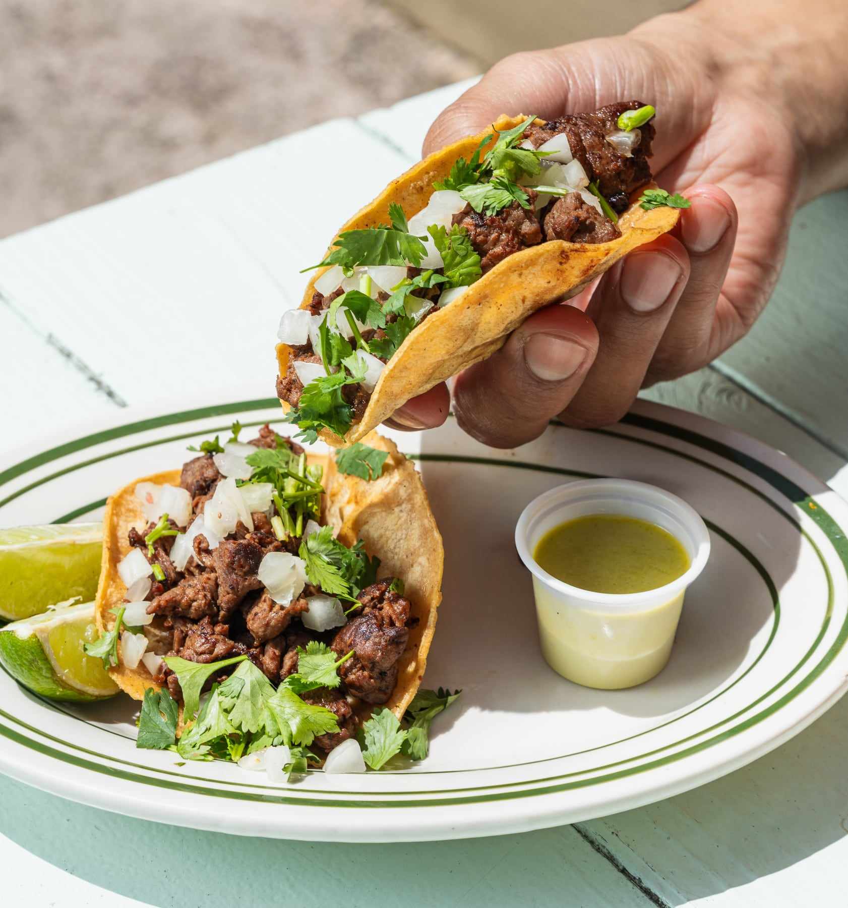 A carne asada taco with onion and cilantro held over a plate, with a cup of green salsa