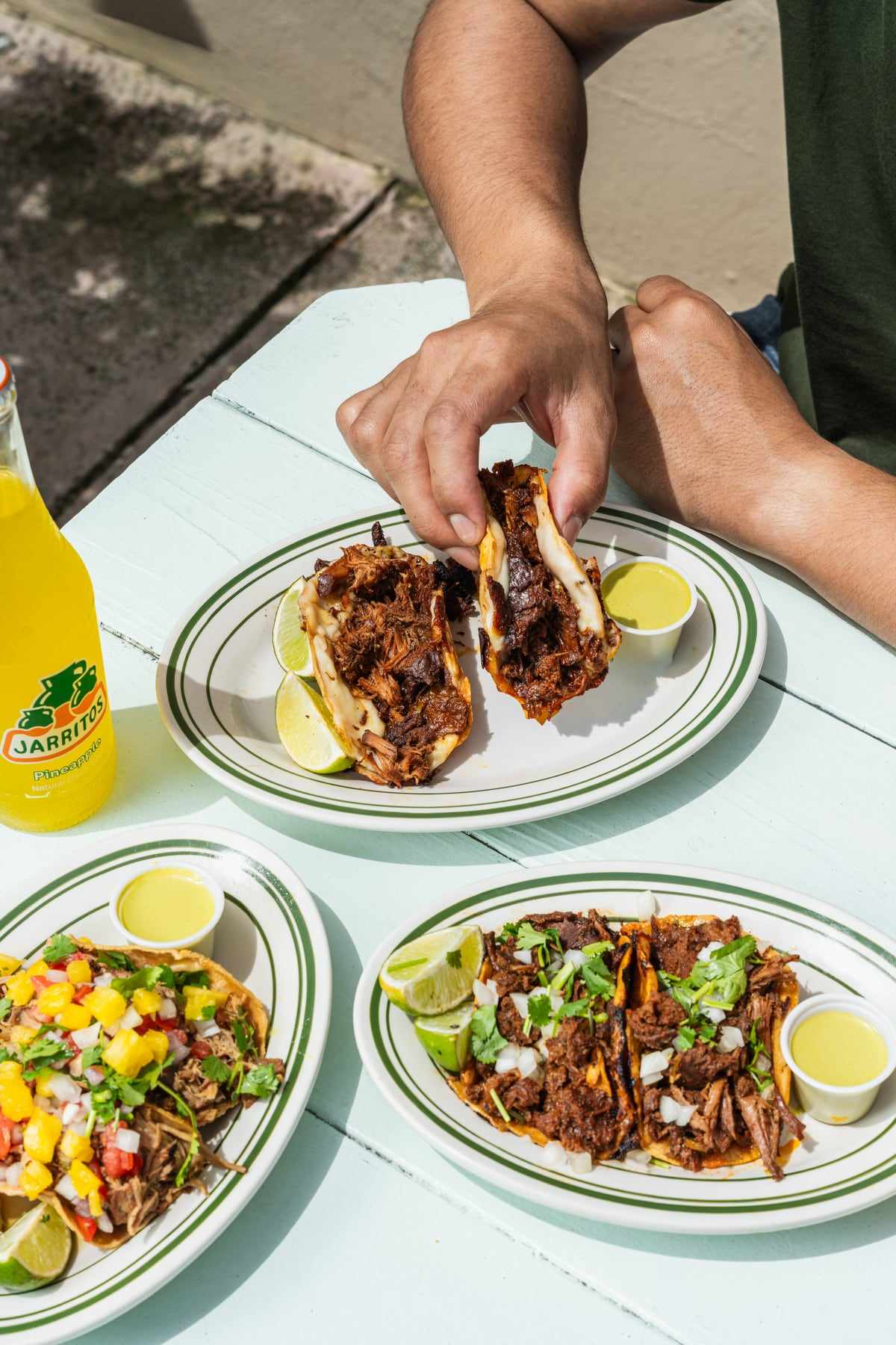 Two birria tacos held over a plate of street tacos, with a bottle of Jarritos on the table